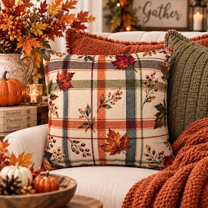 May include: A square throw pillow featuring a cream-colored base with a plaid pattern in shades of orange, green, and dark blue. The design is decorated with printed autumn maple leaves and clusters of red berries scattered across the fabric surface.