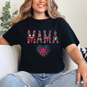 Peut inclure: T-shirt noir avec le mot "MAMA" dans un motif floral. En dessous du texte, un motif floral en forme de cœur. Le t-shirt est fait d'une matière douce.