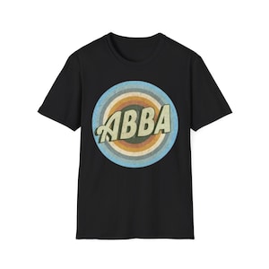 Pode incluir: Uma t-shirt preta de manga curta com um gráfico circular no peito. O design inclui a palavra ABBA numa fonte clara e estilizada, centrada dentro de anéis concêntricos de azul, laranja e castanho com uma textura de aspeto desgastado.