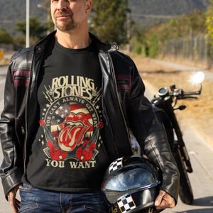 Peut inclure: T-shirt noir avec un graphisme des Rolling Stones représentant le logo de la langue du groupe, des guitares et le texte "You Can't Always Get What You Want". La personne porte une veste en cuir noire et tient un casque de moto à damier.
