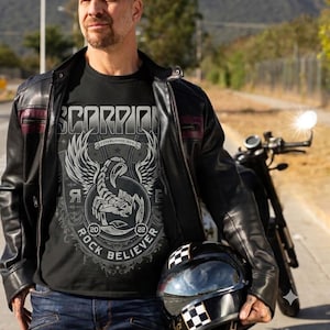 Könnte beinhalten: Ein schwarzes T-Shirt mit einer Grafik eines geflügelten Skorpions, dem Text SCORPION, ESTABLISHED 1985, 20, 22 und ROCK BELIEVER. Das T-Shirt wird unter einer schwarzen Lederjacke mit weinroten Akzenten, blauen Jeans und einem schwarzen Helm getragen.