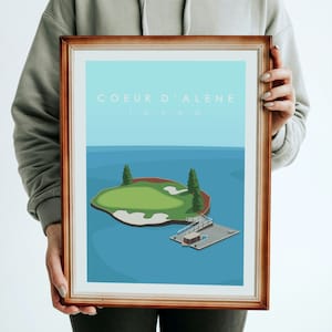 Puede incluir: Una ilustración enmarcada de un campo de golf en una isla en Coeur d'Alene, Idaho. La ilustración es de estilo minimalista con un fondo azul y un campo de golf verde.