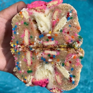 May include: A close-up of a cookie with pink frosting and sprinkles. The cookie is broken in half, revealing a white cream filling and a layer of cookie crumbs.