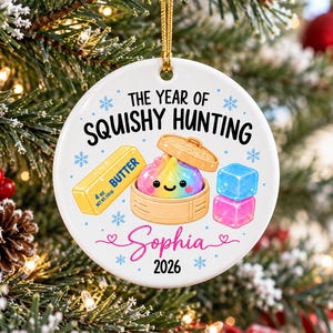 May include: A round white ceramic ornament featuring the text THE YEAR OF SQUISHY HUNTING, Sophia, and 2026. The graphic shows a rainbow-colored dumpling in a bamboo steamer, a yellow stick of butter labeled 4 oz, and two translucent ice cubes.