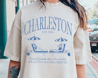 May include: Beige oversized t-shirt with blue text and graphics. The text reads "Charleston Est. 2024" above a graphic of two beach chairs with umbrellas. Below the graphic, it says "Coast Grandmothers Social Club Reagan's Bachelorette".