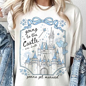 May include: Ivory t-shirt featuring a blue castle graphic with the text "Going to the Castle, gonna get married." The design includes a bow, flowers, and a magic wand. The shirt has a relaxed fit.