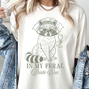 May include: Ivory-colored t-shirt featuring a raccoon wearing heart-shaped sunglasses and holding a wine glass. The raccoon is adorned with a floral crown. The text "IN MY FERAL Bride Era" is printed below the raccoon.