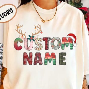 Peut inclure: T-shirt ivoire avec un motif de Noël. Le design comprend les mots "CUSTOM NAME" dans une police à motifs, des bois de rennes et un bonnet de Père Noël. Le t-shirt a une coupe ample.