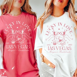 Peut inclure: Deux t-shirts avec un motif graphique rose qui dit "Lucky in Love" avec un verre à martini, des cartes à jouer et des dés. Le texte "Est. 2025 Las Vegas Morgan's Bachelorette" est sous le graphique. Un t-shirt est rose pastèque et l'autre est blanc.