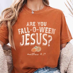 Puede incluir: Camiseta naranja con el texto blanco "ARE YOU FALL-O-WEEN JESUS?", ilustraciones de calabazas y la referencia bíblica Mateo 4:17. La camiseta se combina con pantalones cortos de mezclilla y un cárdigan color crema.