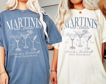 May include: Two oversized t-shirts with a graphic design of martini glasses and the text "MARTINIS and Bikinis" with the text "Amanda's Bachelorette WEST PALM BEACH" and the years "EST. 2024". The t-shirt on the left is blue jean and the t-shirt on the right is ivory.