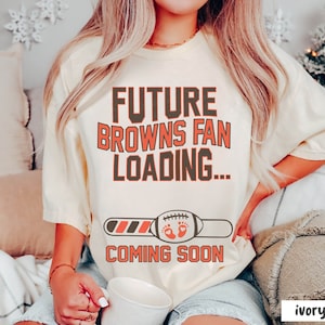 May include: A woman wearing a light yellow t-shirt with the text "FUTURE BROWNS FAN LOADING... COMING SOON" and a progress bar with a football and baby footprints. The shirt is on a white background.