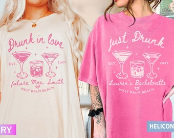 May include: Two t-shirts with a hen party theme. The ivory t-shirt says "Drunk in love" with a cocktail glass and a glass of whisky. The pink t-shirt says "Just Drunk" with two cocktail glasses and a glass of whisky. Both shirts say "EST. 2024" and have a heart with "West Palm Beach" underneath.