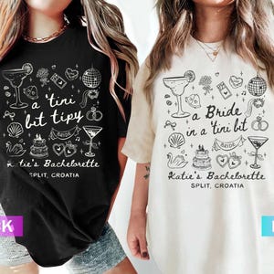 Op de afbeelding: Zwarte en ivoorkleurige T-shirts met een witte grafische opdruk. Het ontwerp bevat cocktailglazen, een discobal en de tekst "a tini bit tipy" en "a Bride in a tini bit". De shirts zijn voor een vrijgezellenfeest in Split, Kroatië.