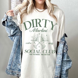 Puede incluir: Camiseta de color marfil con un gráfico verde que dice "DIRTY Martini SOCIAL CLUB". El diseño incluye copas de martini cruzadas y la frase "THE filthier, THE better". La camiseta se combina con una chaqueta vaquera.