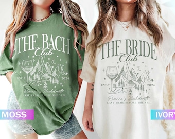 May include: Two t-shirts featuring a mountain and wine glass design. The green shirt reads "The Bach Club" and the white shirt reads "The Bride Club". Both shirts include the text "Racanna's Bachelorette Last Trail Before The Veil" and "Est. 2024".