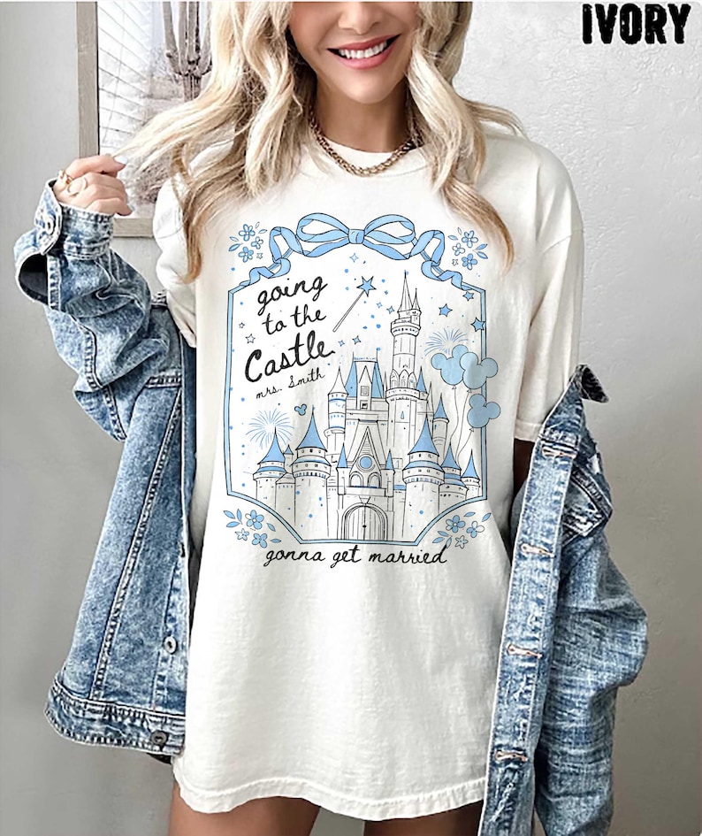 May include: Ivory t-shirt featuring a blue castle graphic with the text "Going to the Castle, gonna get married." The design includes a bow, flowers, and a magic wand. The shirt has a relaxed fit.
