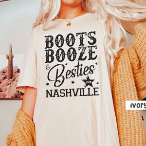 Może przedstawiać: Kremowa koszulka z napisem "Boots, Booze & Besties Nashville 2024" w czarnym, stylizowanym na vintage kroju pisma. Koszulka ma luźny kr&oacute;j i kr&oacute;tkie rękawy. Tekst jest wyśrodkowany na przodzie.