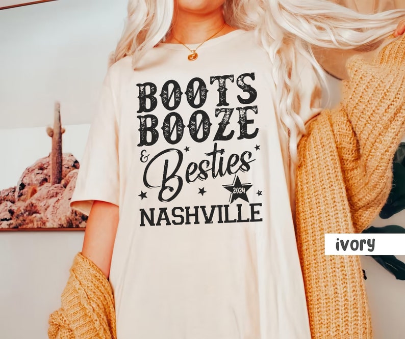 Może przedstawiać: Kremowa koszulka z napisem "Boots, Booze & Besties Nashville 2024" w czarnym, stylizowanym na vintage kroju pisma. Koszulka ma luźny kr&oacute;j i kr&oacute;tkie rękawy. Tekst jest wyśrodkowany na przodzie.