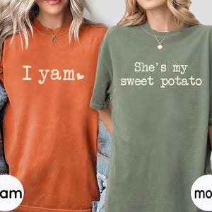 以下が含まれることがあります： 白文字のオレンジとモスグリーンのTシャツ2枚。「I yam」とハートマークがプリントされたオレンジのシャツと、「She's my sweet potato」と書かれたグリーンのシャツ。シャツの下部には「Yam」と「moss」の文字があります。