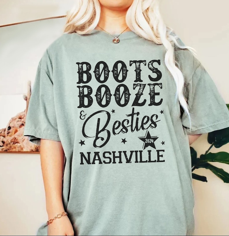 Może przedstawiać: Szałwiowa koszulka z napisem "Boots, Booze & Besties Nashville 2024" w czarnym, postarzanym kroju. Projekt zawiera gwiazdy i grafikę gwiazdy. Koszulka ma kr&oacute;tkie rękawy i luźny kr&oacute;j.