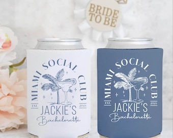 Può includere: Due refrigeratori per lattine bianchi e blu con un design a forma di palma e cocktail. Il testo recita "Miami Social Club Est. 2021 Jackie's Bachelorette". Un refrigeratore è bianco e l'altro è blu.