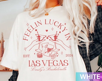 May include: White t-shirt with a red graphic that reads "Feelin' Lucky In Las Vegas" with martini glasses, playing cards, and dice. The text "Emily's Bachelorette" is also included. The year "2024" is printed below the text.