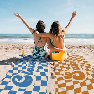 May include: Two people are sitting on beach towels with checkered patterns. The towels have the words "Clover" and "Cream" printed on them in a retro font.