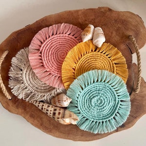 Puede incluir: Un conjunto de posavasos de macramé hechos a mano en varios colores, incluyendo rosa, amarillo y turquesa, dispuestos sobre una bandeja de madera. Los posavasos tienen un diseño circular con un borde con flecos. Varias conchas marinas se colocan alrededor.
