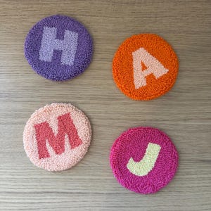 Puede incluir: Cuatro posavasos circulares coloridos con letras bordadas. Los posavasos son morados con una "H" blanca, naranjas con una "A" blanca, melocotón con una "M" roja y rosas con una "J" amarilla. Sobre una superficie de madera marrón claro.