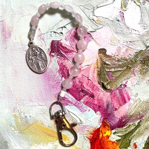 May include: A pink and white beaded rosary with a silver charm featuring a religious image. The rosary is attached to a silver clasp.