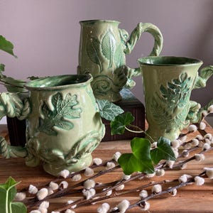 Puede incluir: Tres tazas de cerámica verde con diseños de hojas y vides. Las tazas tienen asas ornamentadas y están decoradas con patrones botánicos detallados. Las tazas se exhiben sobre una superficie de madera con vegetación.