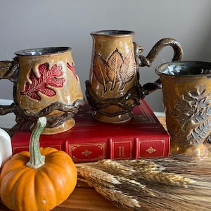 May include: Three ceramic mugs with unique, nature-inspired designs. The mugs feature brown, textured handles and bases, with leaf and branch details. One mug has a red leaf design, another has three brown leaves, and the third has a detailed leaf pattern.