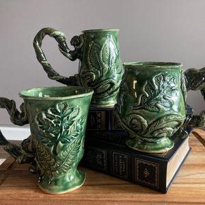 May include: Three green ceramic mugs with leaf and vine designs. The mugs have unique handles shaped like twisted vines. The mugs are different sizes and sit on top of books on a wooden surface.