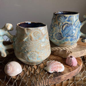 Puede incluir: Dos tazas de cerámica con diseños marinos. Una taza es de color beige con detalles azules, la otra es azul. Ambas tienen asas únicas y están sobre posavasos de madera con conchas marinas y una red de pesca.