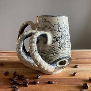 May include: A beige ceramic mug featuring a sculpted octopus tentacle handle and a black ink illustration of a sailing ship on a stormy sea. The mug has a textured surface with visible suction cup details on the tentacles and a dark rim.