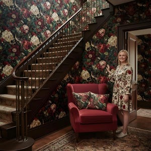 May include: A red armchair with floral pillows sits on a patterned rug in front of a staircase. The walls are covered in dark floral wallpaper. A person wearing a floral dress stands near the door.