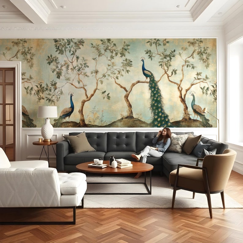 May include: A living room interior with a large wall mural featuring a vintage illustration of peacocks in a forest setting. A gray sectional sofa with pillows is positioned in front of the mural. A white armchair and a round wooden coffee table are also in the room.