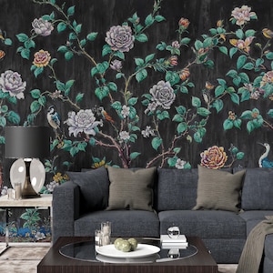 May include: A dark floral wallpaper with large, colourful flowers and green leaves against a black background. The design includes birds perched on branches. A dark grey sofa with cushions and a coffee table are in front of the wallpaper.