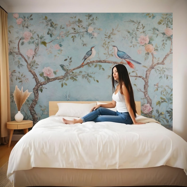 May include: A woman sits on a white bed with a floral patterned wallpaper behind her. The wallpaper features a blue and green tree with pink flowers and blue birds.
