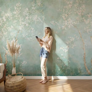 Puede incluir: Una mujer con un su&eacute;ter rosa y pantalones cortos de mezclilla azul se para frente a una pared con un dise&ntilde;o de papel tapiz floral. Ella sostiene un tel&eacute;fono inteligente en su mano y mira hacia un lado.