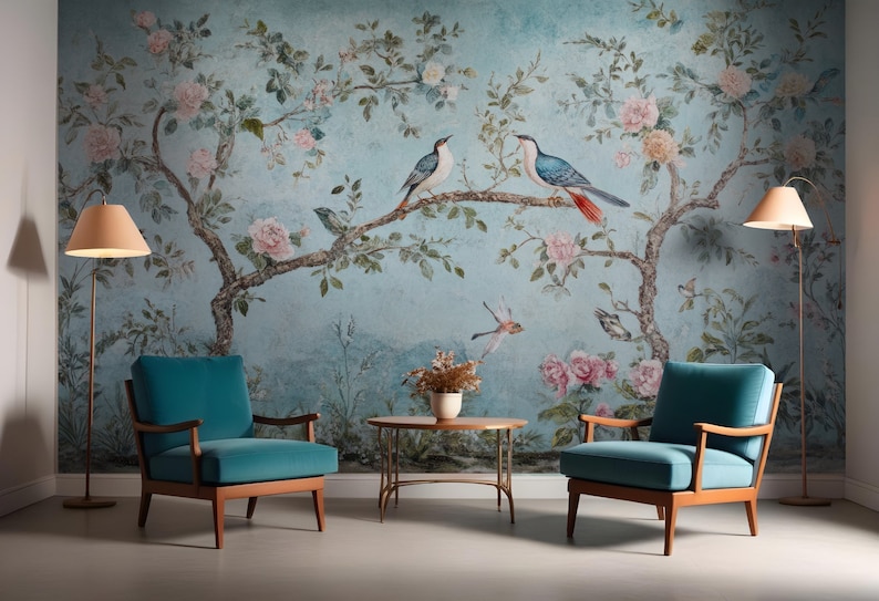 May include: Two teal blue armchairs with wooden frames sit in front of a wall mural depicting a blue and green floral pattern with pink flowers and blue birds. A small round table with a gold metal frame sits between the chairs.