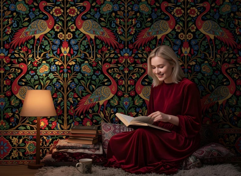 May include: A woman in a red dress reads a book in front of a colorful floral and bird patterned wallpaper. A lamp with a beige shade and a stack of books are visible. The scene is set on a rug with pillows.