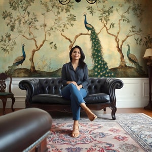 May include: A woman sits on a black leather sofa in front of a wall mural depicting peacocks and trees. The mural is painted in shades of green, blue, and brown. The woman is wearing a gray shirt and blue jeans. She is looking at the camera.