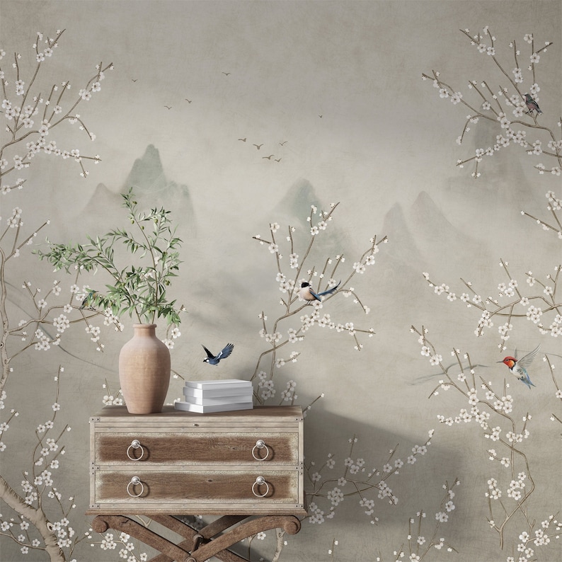Op de afbeelding: Een lichtgrijze muur met een bloemenbehang met witte bloesems en vogels. Een houten bijzettafel met twee laden staat voor de muur. Een vaas met een groene plant staat op de tafel.