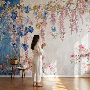 May include: A woman stands before a floral mural featuring cascading pink wisteria, blue butterflies, and lush green foliage. The mural's design includes a blue watercolor section and a small table with a basket of plants. The woman wears a white jumpsuit.