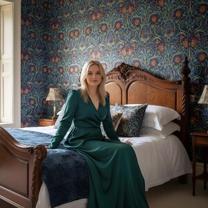 May include: A woman in a long, emerald green dress sits on a bed with a dark wood headboard. The room features patterned wallpaper in blue, orange, and gold tones. The bed has white linens and decorative pillows.