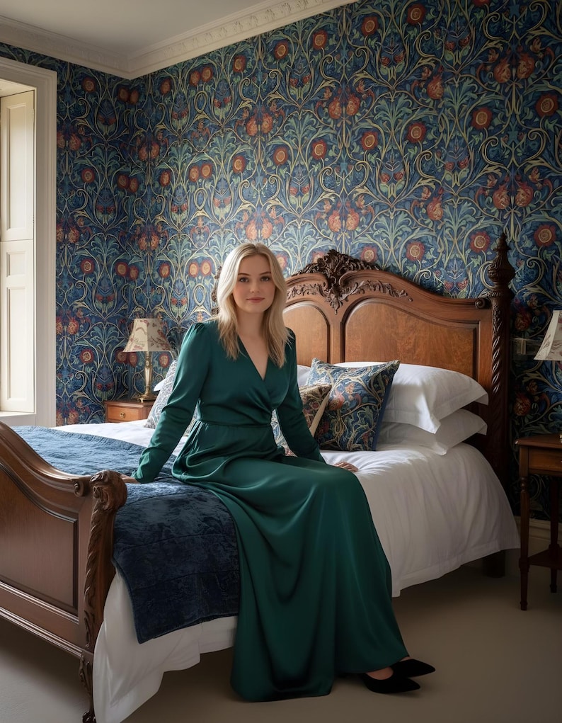 May include: A woman in a long, emerald green dress sits on a bed with a dark wood headboard. The room features patterned wallpaper in blue, orange, and gold tones. The bed has white linens and decorative pillows.