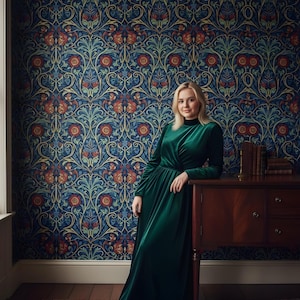 May include: A woman in a long, emerald green velvet dress stands next to a dark wood cabinet. The background features a patterned wallpaper with a blue base and floral designs in red, orange, and teal. A patterned rug is on the floor.