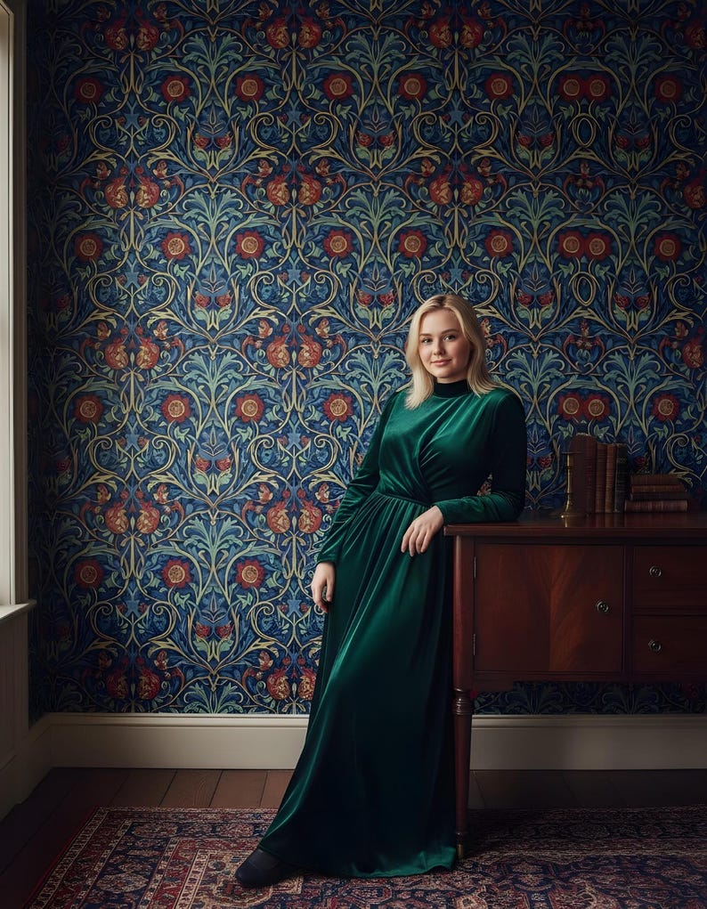 May include: A woman in a long, emerald green velvet dress stands next to a dark wood cabinet. The background features a patterned wallpaper with a blue base and floral designs in red, orange, and teal. A patterned rug is on the floor.